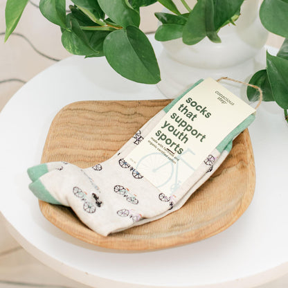 Pair of light green socks featuring bicycles with a packaging label on a wooden tray with a plant in the background