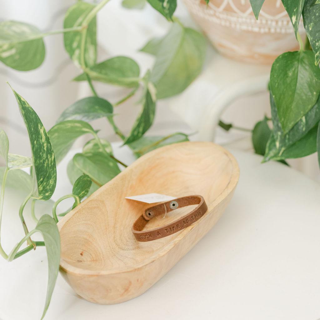Wooden dish with a dark leather bracelet featuring the saying "for such a time as this", surrounded by green leaves on a light background