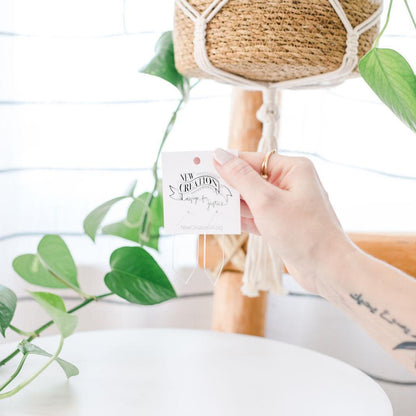 Hand holding a branded card with thin silver hexagon hoops in front of a decorative plant and woven basket.