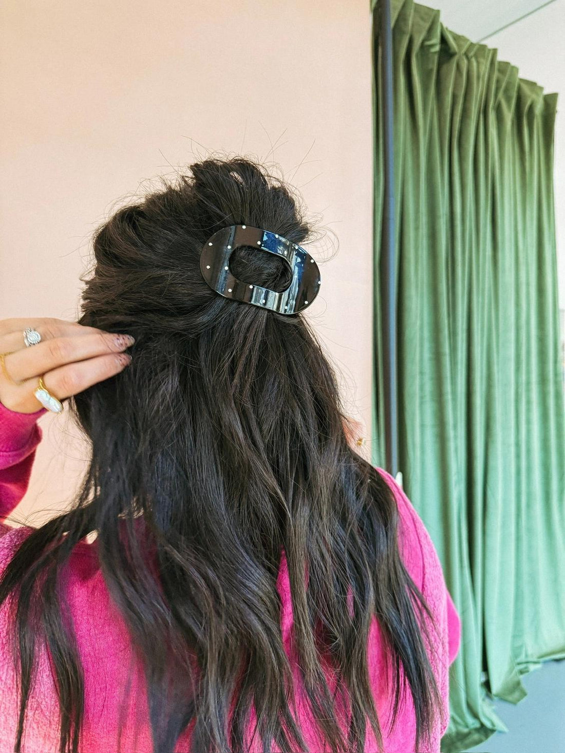 Person with long black hair held back with a black + white hair clip, wearing a pink top, in front of a green curtain.