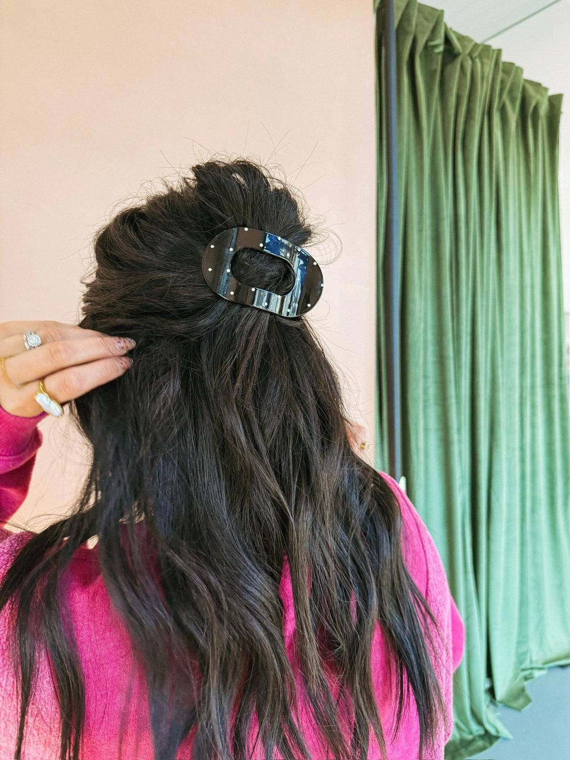 Person with long black hair held back with a black + white hair clip, wearing a pink top, in front of a green curtain.