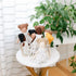 A collection of handcrafted dolls with varying skin tones and hairstyles displayed alongside a brass basket and green plants in the background.