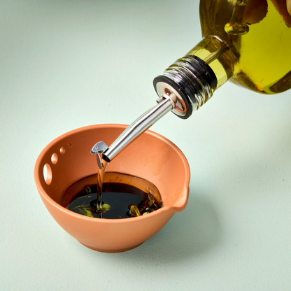 Olive oil being poured from a bottle into a terracotta colored bowl on a light green background