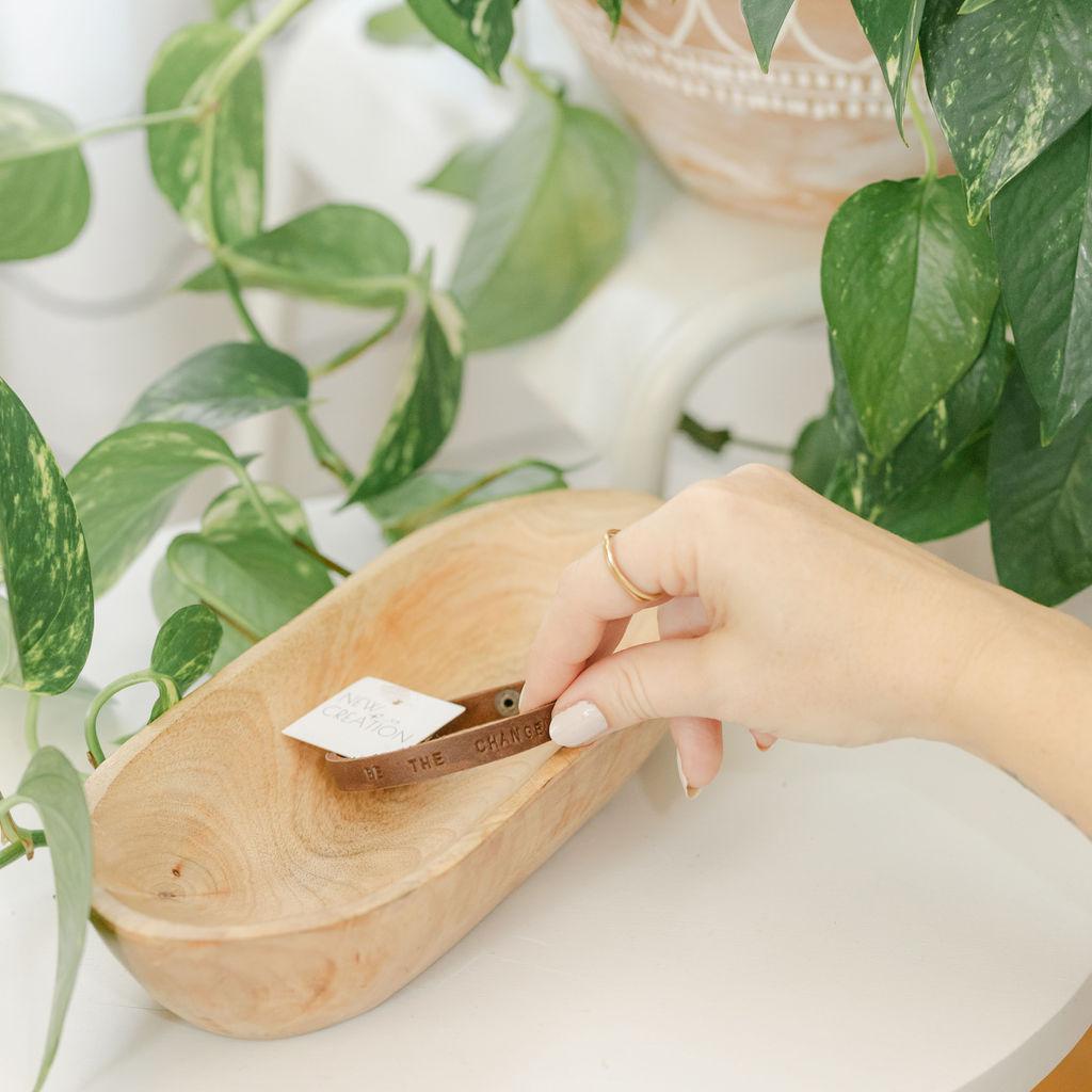 Hand reaching into a wooden dish with a brown leather bracelet with &